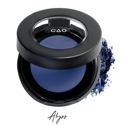 Semi- open eyeshadow compact with blue compressed eyeshadow in shade "Abyss"powder and on white background with loose eyeshadow powder on white..