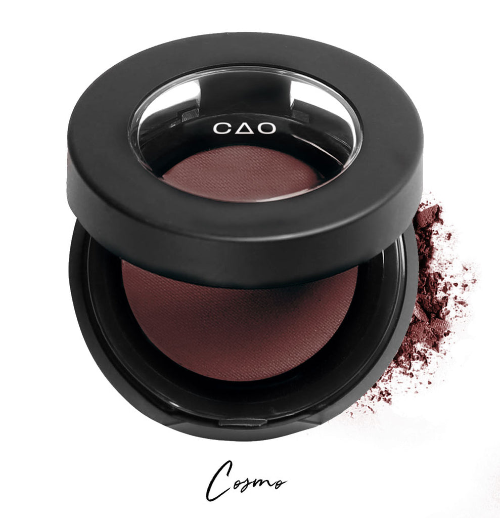 Semi- open eyeshadow compact with dark burgundy eyeshadow in shade "Cosmo" compressed powder and on white background with loose eyeshadow powder on white..