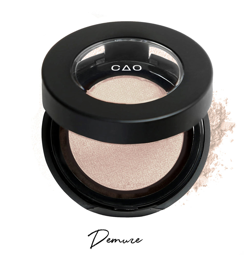 Semi-open eyeshadow compact with biege eyeshadow in shade "demure" compressed powder and on white background with loose eyeshadow powder on white.