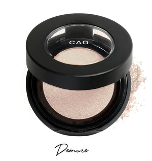 Semi-open eyeshadow compact with biege eyeshadow in shade "demure" compressed powder and on white background with loose eyeshadow powder on white.