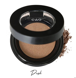Semi- open eyeshadow compact with light brown eyeshadow in shade "Dusk" compressed powder and on white background with loose eyeshadow powder on white..