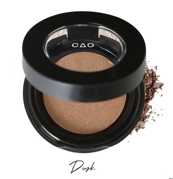 Semi- open eyeshadow compact with light brown eyeshadow in shade "Dusk" compressed powder and on white background with loose eyeshadow powder on white..