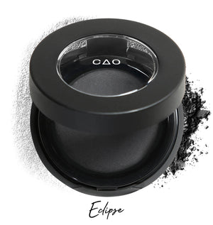 Semi- open eyeshadow compact with black eyeshadow in shade "eclipse" compressed powder and on white background with loose eyeshadow powder on white.