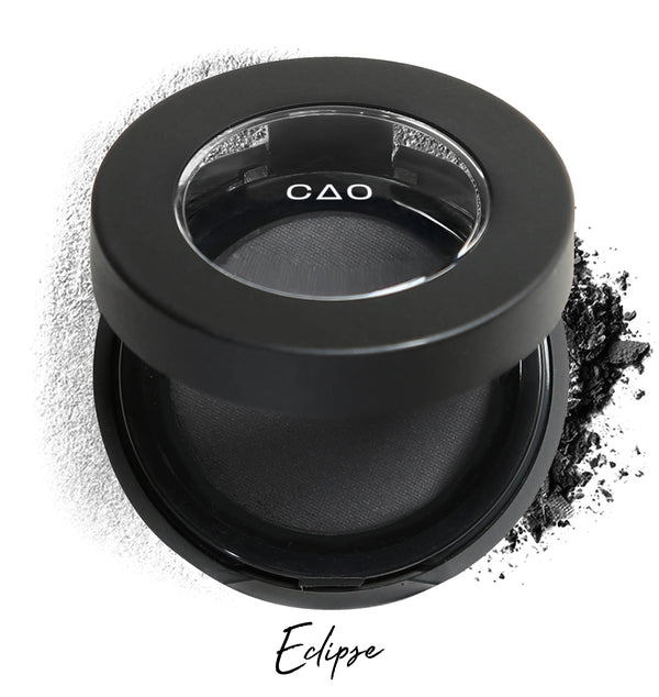 Semi- open eyeshadow compact with black eyeshadow in shade "eclipse" compressed powder and on white background with loose eyeshadow powder on white.