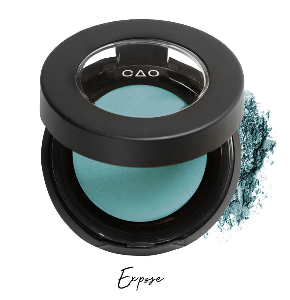 Semi- open eyeshadow compact with teal eyeshadow in shade "expose" compressed powder and on white background with loose eyeshadow powder on white..