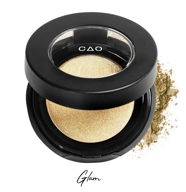 Semi- open eyeshadow compact with light shimmer gold eyeshadow in shade "glam" compressed powder and on white background with loose eyeshadow powder on white..