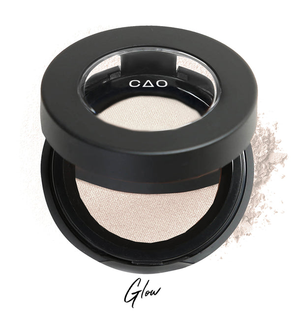 Semi-open eyeshadow compact with white eyeshadow in shade "glow" compressed powder and on white background with loose eyeshadow powder on white.