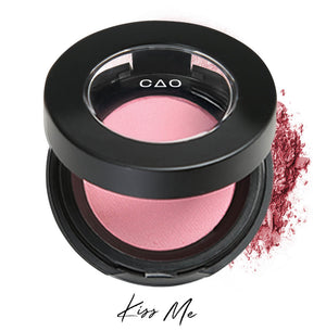 Semi- open eyeshadow compact with light pastel pink eyeshadow in shade "Kiss Me" compressed powder and on white background with loose eyeshadow powder on white..