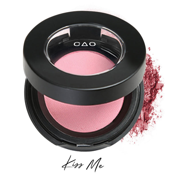 Semi- open eyeshadow compact with light pastel pink eyeshadow in shade "Kiss Me" compressed powder and on white background with loose eyeshadow powder on white..