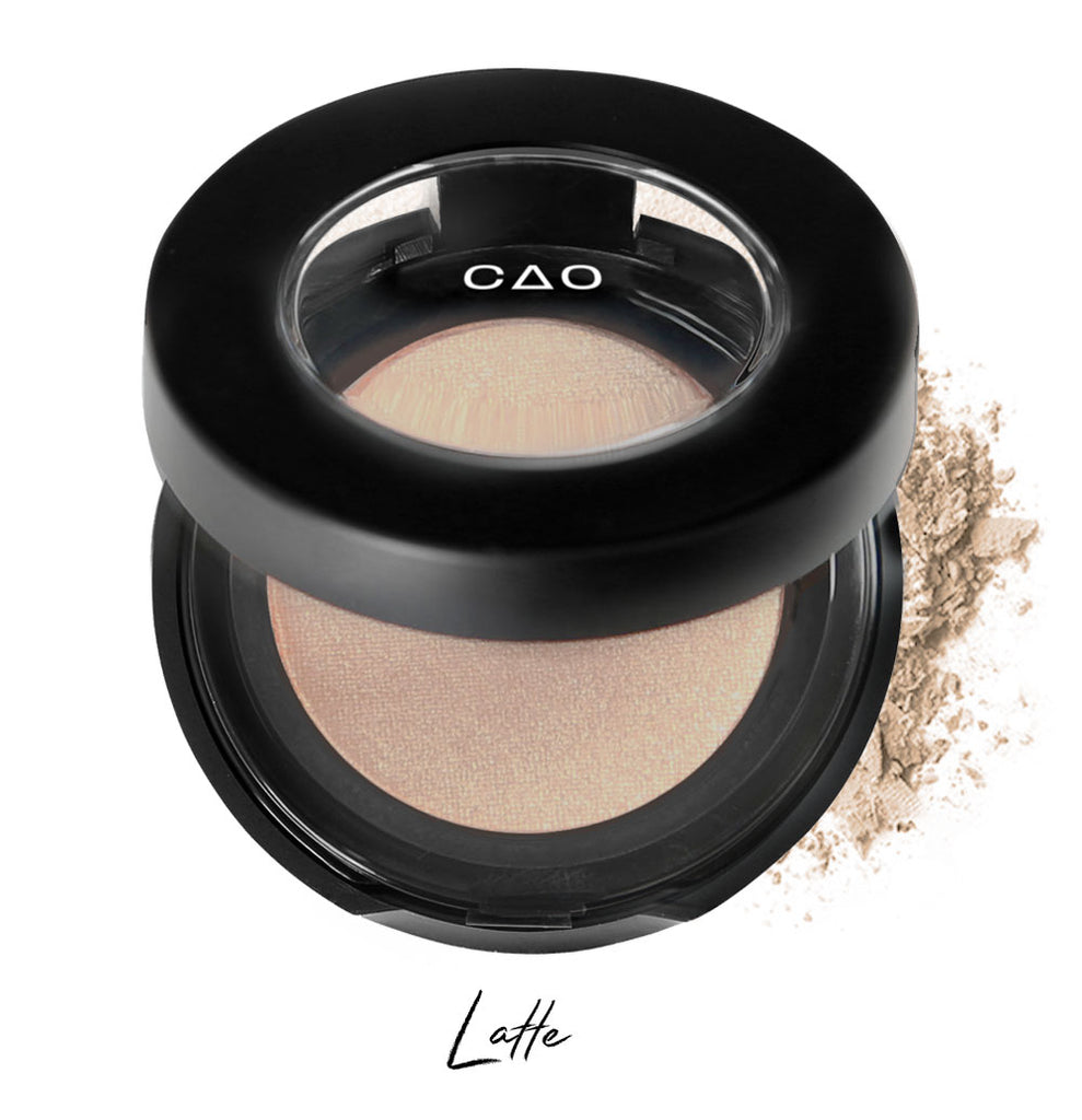 Semi- open eyeshadow compact with light nude eyeshadow in shade "latte" compressed powder and on white background with loose eyeshadow powder on white..