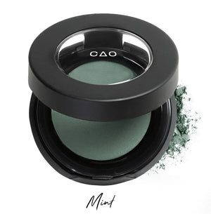 Semi- open eyeshadow compact with desaturated green eyeshadow in shade "mint" compressed powder and on white background with loose eyeshadow powder on white..