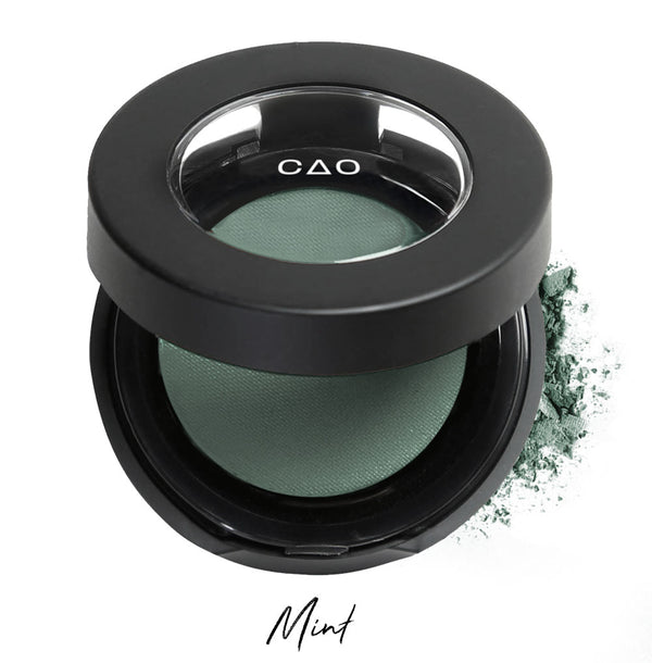 Semi- open eyeshadow compact with desaturated green eyeshadow in shade "mint" compressed powder and on white background with loose eyeshadow powder on white..