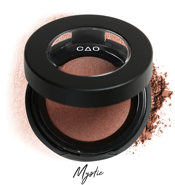 Semi-open eyeshadow compact with warm brown eyeshadow in shade "mystic" compressed powder and on white background with loose eyeshadow powder on white.