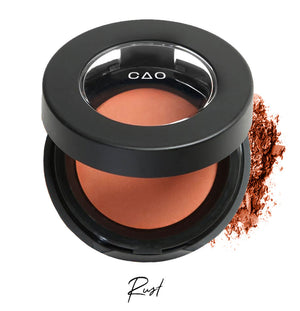 Semi- open eyeshadow compact with rust colored eyeshadow in shade "rust" compressed powder and on white background with loose eyeshadow powder on white..