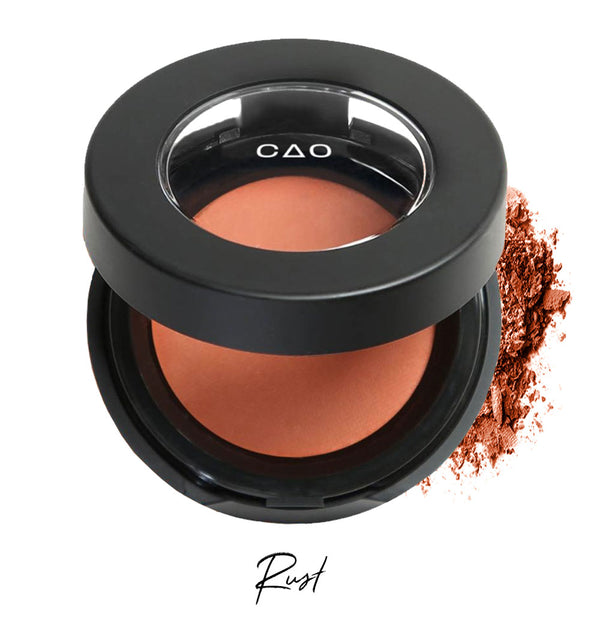 Semi- open eyeshadow compact with rust colored eyeshadow in shade "rust" compressed powder and on white background with loose eyeshadow powder on white..