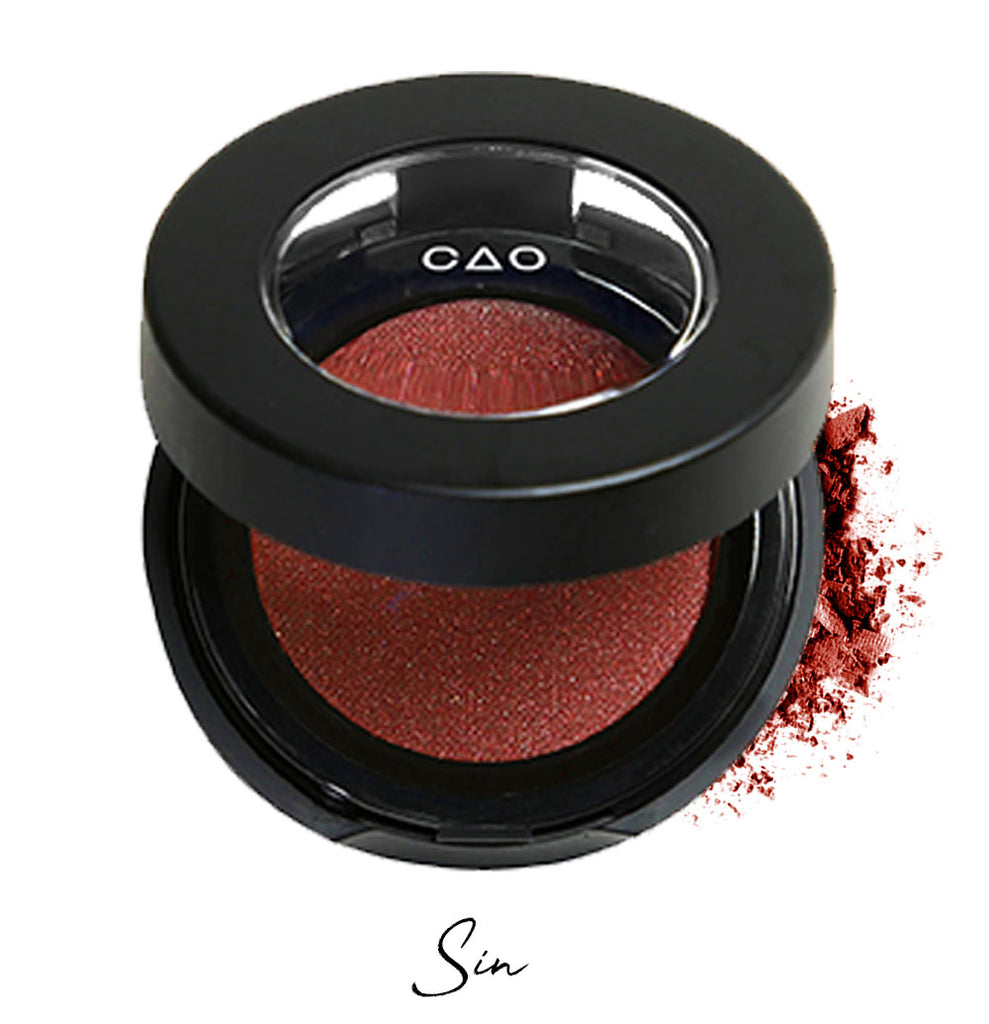Semi- open eyeshadow compact with dark red eyeshadow in shade "sin" compressed powder and on white background with loose eyeshadow powder on white..