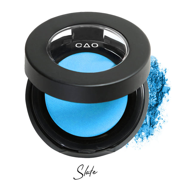 Semi- open eyeshadow compact with light neon blue eyeshadow in shade "Slate" compressed powder and on white background with loose eyeshadow powder on white..