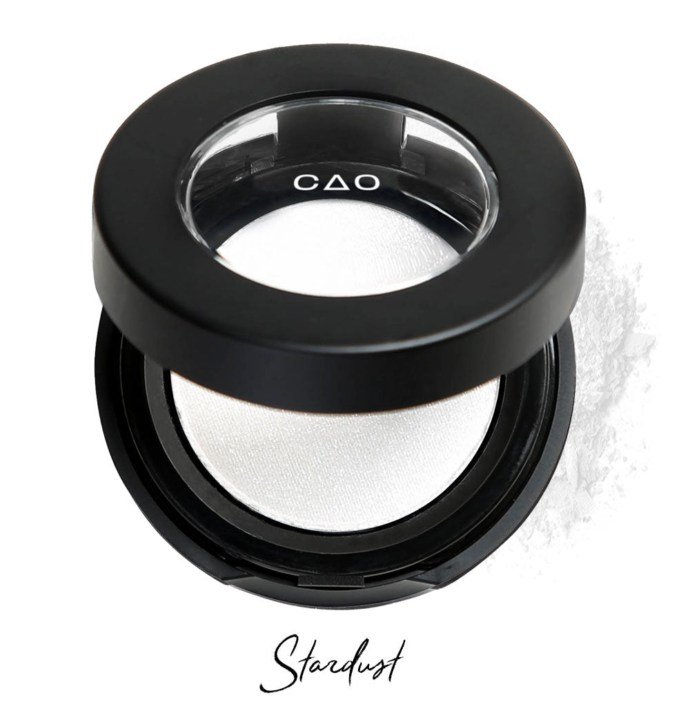 Semi- open eyeshadow compact with white shimmer eyeshadow in shade "stardust" compressed powder and on white background with loose eyeshadow powder on white..