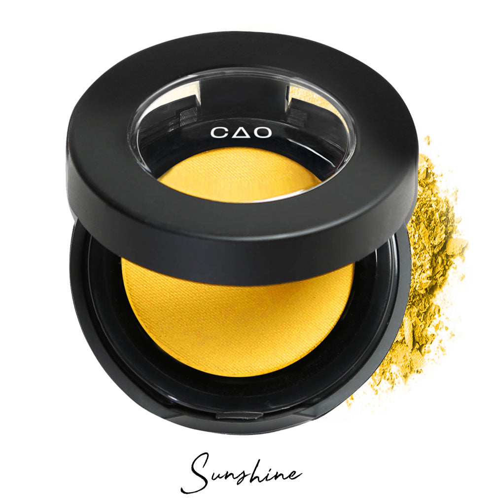 Semi- open eyeshadow compact with yellow eyeshadow in shade "sunshine" compressed powder and on white background with loose eyeshadow powder on white..