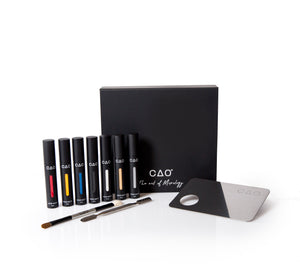 6 Black Matted tubes, vertical stretched oval windows, each filled filled with blue, red, yellow, silver metallic, gold metallic, white, and black liquid matte makeup. Closed black liquid matte collection box, next to metal mixing palette and spatula along with double ended brush white bristle and brown bristle. 