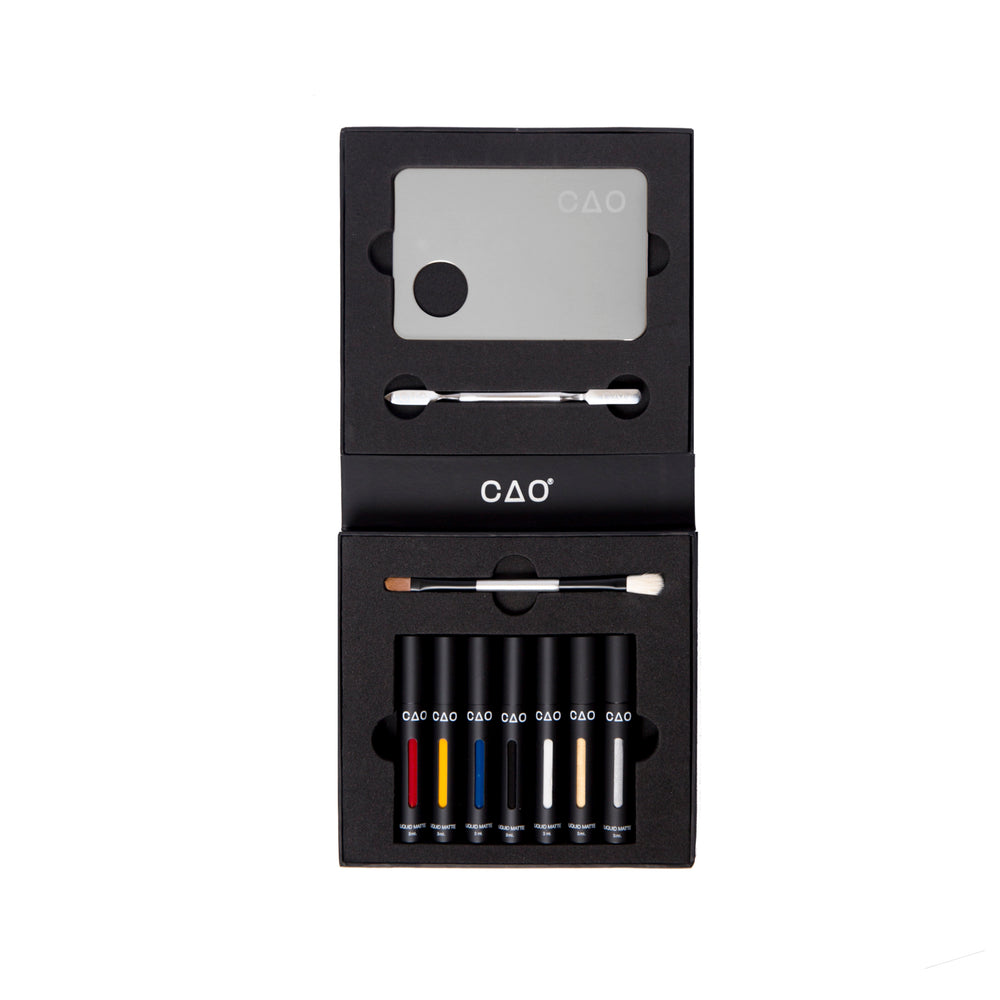 6 Black Matted tubes, vertical stretched oval windows, each filled filled with blue, red, yellow, silver metallic, gold metallic, white, and black liquid matte makeup. Closed black liquid matte collection box, next to metal mixing palette and spatula along with double ended brush white bristle and brown bristle. 