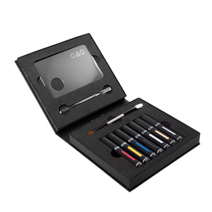 opened black liquid matte collection box, top side metal mixing palette and spatula, bottom double ended brush white bristle and brown bristle. 6 Black Matted tubes, vertical stretched oval windows, each filled filled with blue, red, yellow, silver metallic, gold metallic, white, and black liquid matte makeup.