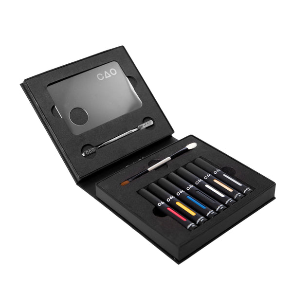 opened black liquid matte collection box, top side metal mixing palette and spatula, bottom double ended brush white bristle and brown bristle. 6 Black Matted tubes, vertical stretched oval windows, each filled filled with blue, red, yellow, silver metallic, gold metallic, white, and black liquid matte makeup.
