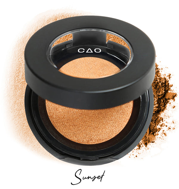 Semi- open eyeshadow compact with light orange eyeshadow in shade "sunset" compressed powder and on white background with loose eyeshadow powder on white.