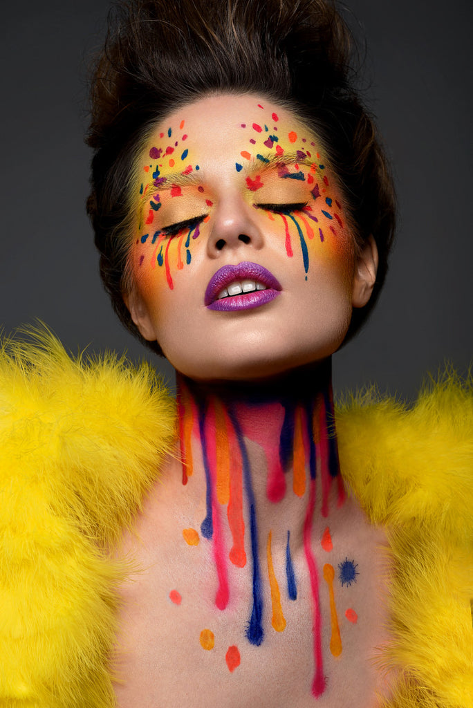 Model with airbrush makeup on closed eyes and neck, splattered and dripping colors orange, red, blue, purple, on yellow. Purple lip with pink center. Brown hair pulled back from face. Model wearing yellow fur bolero on dark gray back ground.